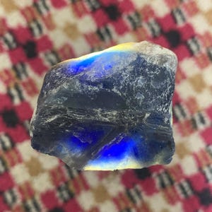 Könnte beinhalten: Ein roher, unpolierter Edelstein mit einem tiefblauen, durchscheinenden Kern, der in intensiver Farbe leuchtet. Die Außenseite weist eine raue, strukturierte Oberfläche mit grauen und weißen mineralischen Einschlüssen auf.