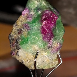 Puede incluir: Primer plano de una gema en bruto, que muestra una vibrante mezcla de colores. La piedra presenta un tono magenta intenso, intercalado con áreas de verde brillante y motas de tonos más claros. Se sujeta con un soporte de metal plateado.