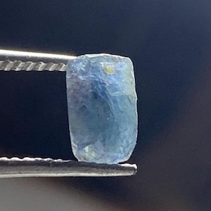 Könnte beinhalten: Ein roher, durchscheinender hellblauer Edelstein mit rechteckiger, leicht unregelmäßiger Form und facettierten Kanten. Der Stein zeigt Variationen in der Farbintensität, die von blassem Himmelblau bis zu tieferen Azurtönen reichen und von einer Pinzette gehalten werden.