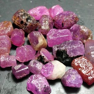 May include: A collection of rough-cut, natural ruby gemstones in various shades of pink and red. The gemstones have a crystalline structure and range in size, with some showing dark inclusions.