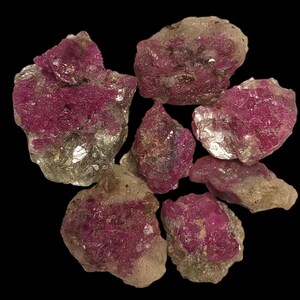 May include: A collection of six raw, unpolished ruby crystals featuring deep magenta and pink hues embedded within translucent, light-colored mineral matrix rock. The stones exhibit irregular, jagged shapes with a mix of opaque and crystalline textures.