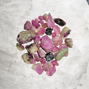 Könnte beinhalten: Eine Sammlung roher Rubin-Edelsteine in verschiedenen Größen und Farbtönen von Rosa, Rot und Grün. Die rauen, ungeschliffenen Steine sind auf einer hellgrauen Oberfläche verstreut und zeigen ihre natürlichen Texturen und Farben. Einige Steine haben ein kristallines Aussehen.