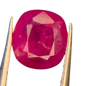 Può includere: Una gemma di rubino di forma ovale con una superficie sfaccettata, che mostra un colore rosso intenso e inclusioni interne. La gemma è tenuta saldamente tra le due punte metalliche di una pinzetta color argento su uno sfondo bianco.