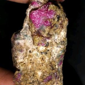 Pode incluir: Uma gema bruta, não lapidada, com um vibrante tom magenta. A pedra está embutida numa matriz de rocha bege e castanha, com estruturas cristalinas visíveis. A aparência geral sugere um espécime mineral natural e não polido.