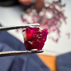 May include: A raw, deep red gemstone held by metal tweezers. The stone features an irregular, multifaceted surface with translucent sections and a small patch of white mineral inclusion on the left side, showcasing a natural, unpolished crystalline texture.