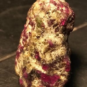 Puede incluir: Una gema en bruto sin cortar con una mezcla de colores. La piedra tiene una base beige con parches de rojo intenso y motas de verde y dorado. La forma general es irregular y texturizada.
