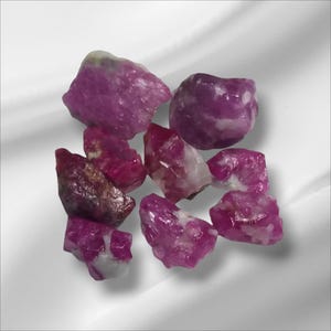 Puede incluir: Un conjunto de piedras preciosas de rubí en bruto. Las piedras, de varios tamaños y formas, muestran un tono rojo-rosado intenso con algunas inclusiones blancas y marrones. Las piedras preciosas están talladas en bruto, mostrando su estado natural sin pulir.