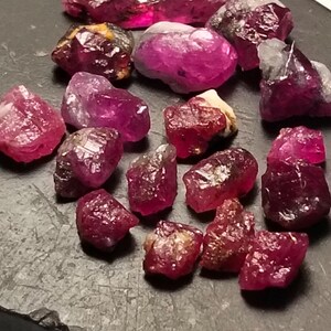 Puede incluir: Una colección de gemas de rubí en bruto de varias formas y tamaños. Los rubíes muestran una gama de tonos rojos y rosas, algunos con apariencia translúcida. Están dispuestas sobre una superficie oscura y texturizada.