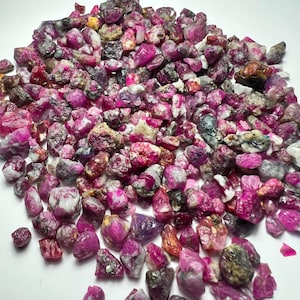 Op de afbeelding: Een verzameling ruwe, ongeslepen robijn-edelstenen in diverse tinten donkerroze, magenta en rood. De stenen hebben onregelmatige, grillige vormen met zichtbare witte, grijze en zwarte minerale insluitingen verspreid over de hele hoop.