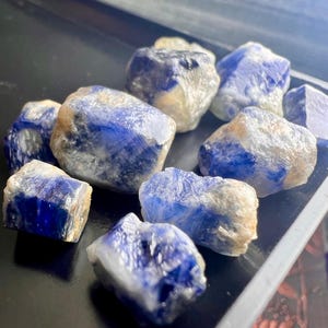 Puede incluir: Una colección de zafiros en bruto, sin cortar. Las piedras preciosas muestran un color azul profundo con áreas más claras, casi blancas. La textura rugosa de las piedras es visible, dispuestas sobre una superficie oscura.