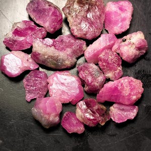 May include: A collection of raw, uncut ruby gemstones. The stones vary in size and shape, displaying a range of pink and red hues. Some have a crystalline structure, while others appear more opaque. The gemstones are scattered on a dark surface.