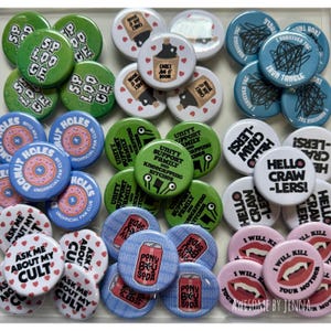 May include: A collection of circular pin-back buttons featuring various graphic designs, including text such as Splooge, Cark's Jug O' Boon, Iron Tangle, Donut Holes, Unity Support Family, Hello Crawlers, Ask Me About My Cult, Pony Baby Soda, and I Will Kill Your Mother.