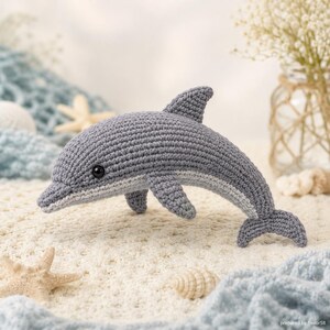 May include: A handcrafted grey crochet dolphin with a white belly stripe and black eyes. The dolphin is displayed on a sandy surface with starfish and seashells, creating a beach theme. The dolphin is a soft toy.