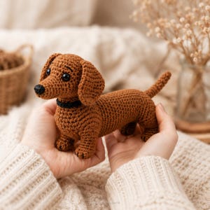Pode incluir: Um cão dachshund de crochê em tons de marrom, com uma coleira preta e um nariz preto. O cão feito à mão é segurado nas mãos de uma pessoa, mostrando o trabalho detalhado de crochê. O corpo do cão é alongado, com pernas pequenas e uma cauda curvada.