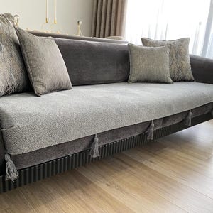 May include: A grey velvet sofa with decorative cushions. The sofa features a textured seat cover and decorative tassels. The legs are gold-coloured. The cushions have various patterns and textures.