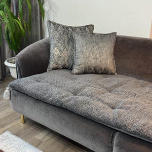 Puede incluir: Un sofá de terciopelo gris con dos cojines decorativos. Un cojín tiene un patrón geométrico con detalles dorados, y el otro tiene una apariencia texturizada y metálica. El sofá tiene un asiento con botones y patas doradas.