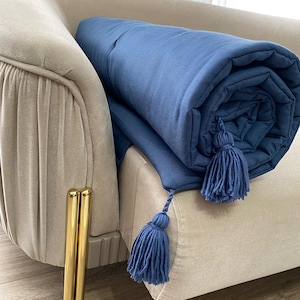 May include: A rolled-up, navy blue blanket with two matching tassels rests on a beige armchair. The blanket is made of a soft material and has a quilted texture. The tassels are at the end of the blanket.