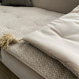 Può includere: Primo piano di una coperta beige con una trama strutturata e un cuscino color crema. Una grande nappa color crema pende dal bordo della coperta. La coperta e il cuscino sono su un divano colorato chiaro.