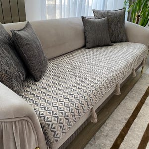 May include: A beige sofa with several gray throw pillows. The sofa cushion has a geometric pattern in shades of gray and beige. Tassels hang from the front of the cushion. The sofa is placed on a light-colored rug.