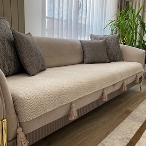 Op de afbeelding: Een beige bank met verschillende grijze kussens. De bank heeft een getextureerde, lichtgekleurde bekleding en is versierd met kwastjes langs de onderrand. Goudkleurige poten geven het meubel een vleugje elegantie.