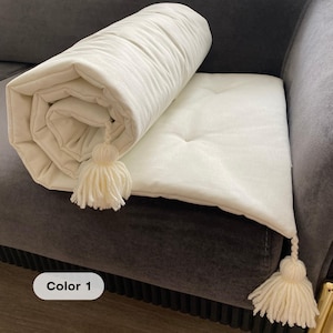 Puede incluir: Un cojín y una esterilla enrollada de color crema con borlas. La esterilla está enrollada y colocada encima del cojín. El cojín tiene un botón en el centro. El texto "Color 1" está en la esquina inferior izquierda.