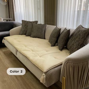May include: A beige sofa with several decorative throw pillows in shades of gray and brown. The sofa has a quilted, cream-colored cushion and decorative tassels. The text "Color 2" is visible on the lower left.