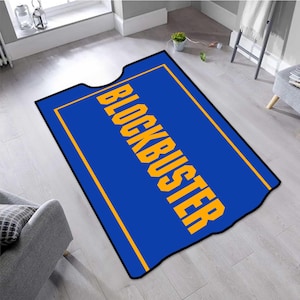 May include: A blue rectangular rug featuring the word "BLOCKBUSTER" in yellow. The rug has a black border and a yellow outline. The rug is placed on a light wooden floor. The rug's design is reminiscent of a movie ticket.