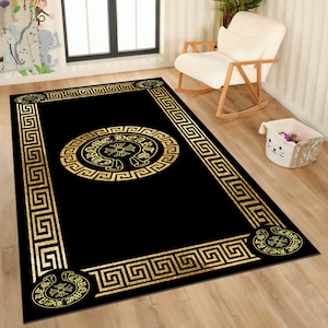 May include: Black rectangular area rug with a gold Greek key border and a central gold medallion design. The medallion features a circular pattern with text and a cross-like emblem. The rug is placed on a light wood floor.