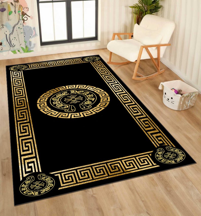 May include: Black rectangular area rug with a gold Greek key border and a central gold medallion design. The medallion features a circular pattern with text and a cross-like emblem. The rug is placed on a light wood floor.