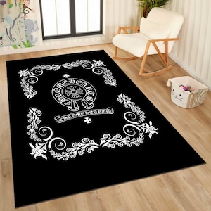 May include: Black rectangular rug with a white floral border and a central emblem. The emblem features the text "CHROME HEARTS" in a circular design, with a cross symbol. The rug is placed on a wooden floor.