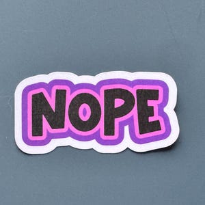 May include: A sticker with the word "NOPE" in black, outlined in pink and purple, and a white border. The sticker is on a gray background.