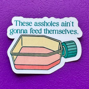 May include: A sticker with the text "These assholes ain't gonna feed themselves." The sticker features a cartoon illustration of a cell culture flask with a pink liquid inside. The flask has a yellow top and a teal cap.