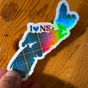 May include: A holographic sticker in the shape of Nova Scotia, with the text "I ♥ NS". The sticker features a plaid pattern in shades of blue and green, with a rainbow gradient effect. The sticker is held by a hand.