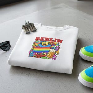 Könnte beinhalten: Weißes Sweatshirt mit einem farbenfrohen „BERLIN“-Grafikdesign mit einem Bären, Regenbogen, Bier und Wurst. Eine schwarze Sonnenbrille und ein Miniatur-Brandenburger Tor liegen neben dem Shirt. Bunt gefärbte Sneaker mit blauen, grünen und rosa Akzenten sind ebenfalls im Bild.