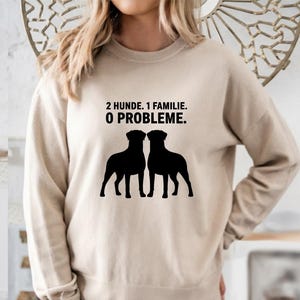 May include: A beige sweatshirt featuring the text "2 Hunde. 1 Familie. 0 Probleme." and a black silhouette of two dogs. The sweatshirt has a crew neck and a relaxed fit.
