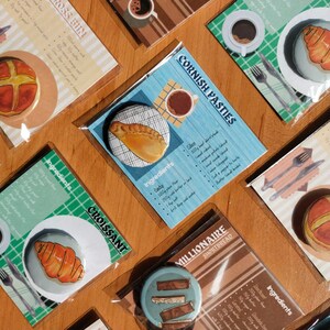 May include: A collection of recipe cards and food-themed badges are arranged on a wooden surface. The cards showcase illustrations of a croissant, Cornish pasty, and hot cross bun, with ingredient lists. The badges feature the same foods, plus millionaire shortbread. The color scheme includes browns, blues, and greens.