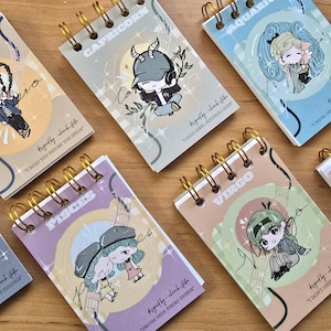 Può includere: Una collezione di piccoli quaderni rilegati a spirale con illustrazioni dei segni zodiacali. Ogni quaderno ha una diversa combinazione di colori pastello e una rilegatura a spirale dorata. I quaderni presentano personaggi dei cartoni animati e testo scritto a mano.