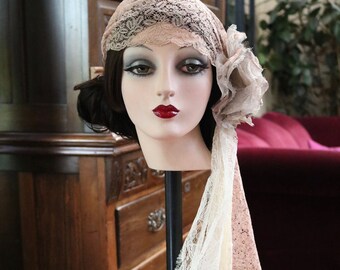 May include: A vintage-inspired lace headpiece in shades of blush and ivory. The headpiece features a delicate floral design and a long, flowing veil.
