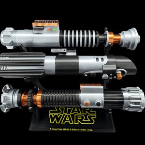 May include: Three Star Wars lightsabers are displayed on a black stand. The lightsabers have silver, black, and orange accents. The stand has the Star Wars logo and the text "A long time ago in a galaxy far, far away."