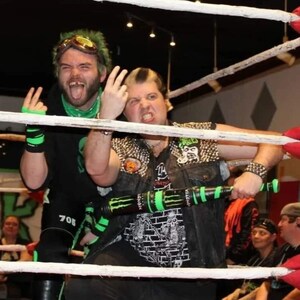 May include: Two men in a wrestling ring, one wearing a green and black wrestling outfit and the other wearing a black vest with silver studs and a black and white graphic t-shirt. The man in the vest is holding a green and black baseball bat with the word "Monster" on it.