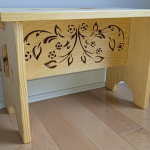 Könnte beinhalten: Ein hellfarbiger Holztritt mit einem floralen Design, das in die Frontplatte geschnitten ist. Der Hocker hat vier Beine und eine flache Oberseite. Das florale Design umfasst Blätter und Blumen. Der Hocker ist aus unfertigem Holz gefertigt.