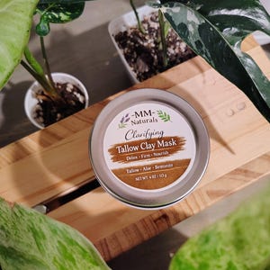 May include: A round, silver-colored tin of "MM Naturals Clarifying Tallow Clay Mask" sits on a wooden surface, surrounded by green plants. The label features text and a brown band with the words "Detox, Firm, Nourish". The net weight is 4 oz / 113 g.