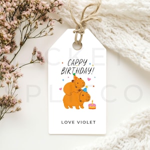 Könnte beinhalten: Weißes Geschenk-Etikett mit Schnur, mit einer Cartoon-Illustration von zwei Capybaras mit Partyhüten. Das Etikett lautet "Cappy Birthday!" und "Love Violet", mit einem kleinen Kuchen. Ideal für ein Geburtstagsgeschenk.