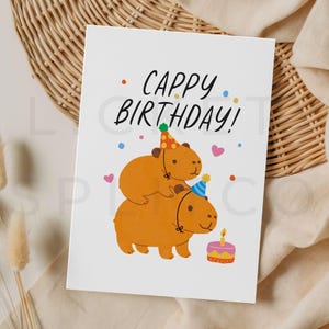 May include: A white greeting card with the text "Cappy Birthday!" features two cartoon capybaras wearing party hats. One capybara is stacked on top of the other. A small cake with a lit candle is next to the bottom capybara.