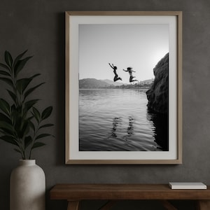Może przedstawiać: Czarno-biała oprawiona fotografia dwóch osób skaczących do wody. Obraz jest osadzony na tle gór i jasnego nieba. Fotografia jest wyświetlana na ścianie nad drewnianą ławką.