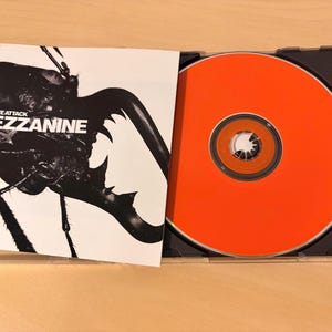 Puede incluir: Un CD de música en una caja de plástico con la portada abierta. La portada presenta un gráfico en blanco y negro de un escarabajo con el texto Massive Attack Mezzanine. El disco es de un color naranja brillante con un orificio central.