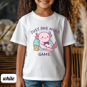 Op de afbeelding: Wit T-shirt met een cartoon axolotl die een videogame speelt. De roze axolotl houdt een zwarte controller vast, met de tekst "JUST ONE MORE GAME" in paars. Een rode drank en een snack zijn ook afgebeeld.