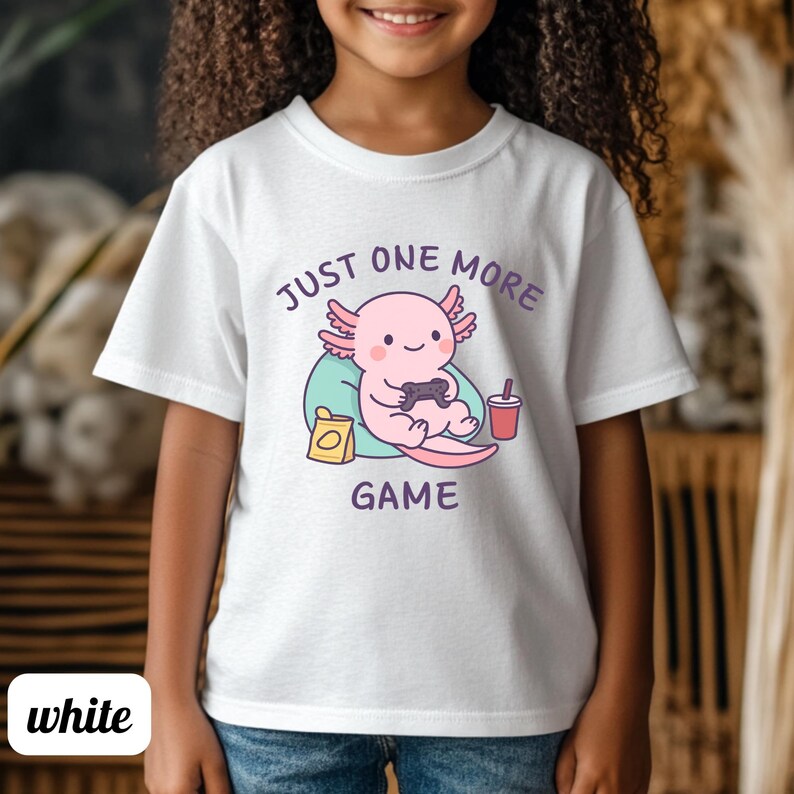 Op de afbeelding: Wit T-shirt met een cartoon axolotl die een videogame speelt. De roze axolotl houdt een zwarte controller vast, met de tekst "JUST ONE MORE GAME" in paars. Een rode drank en een snack zijn ook afgebeeld.