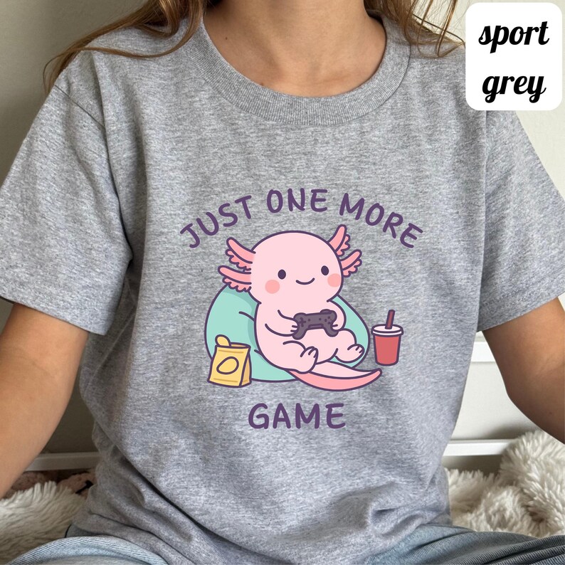 Op de afbeelding: Sportgrijs t-shirt met een cartoon axolotl die een videogame speelt. De roze axolotl houdt een controller vast, met de tekst "JUST ONE MORE GAME". Een drankje en een snack zijn ook afgebeeld.