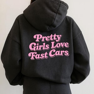 May include: Dark gray hoodie and sweatpants set. The back of the hoodie features the pink text "Pretty Girls Love Fast Cars". The hoodie has a hood and long sleeves. The sweatpants are visible at the bottom of the image.